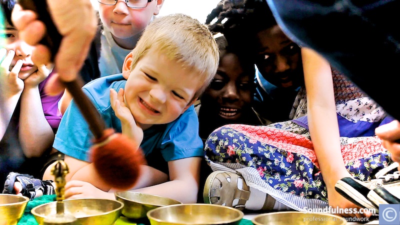 Young child smiling at a row of small singing bowls being played with a mallet