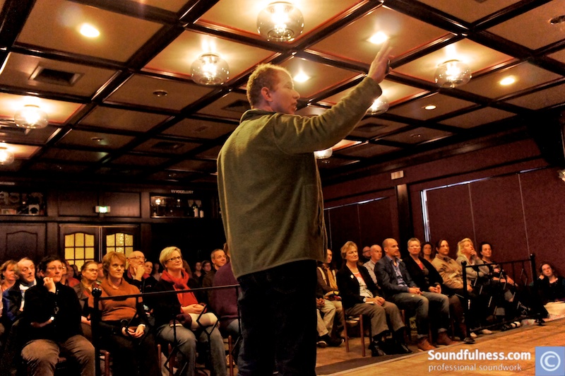 Coen speaking with a raised hand to a seated audience in a warm theatre