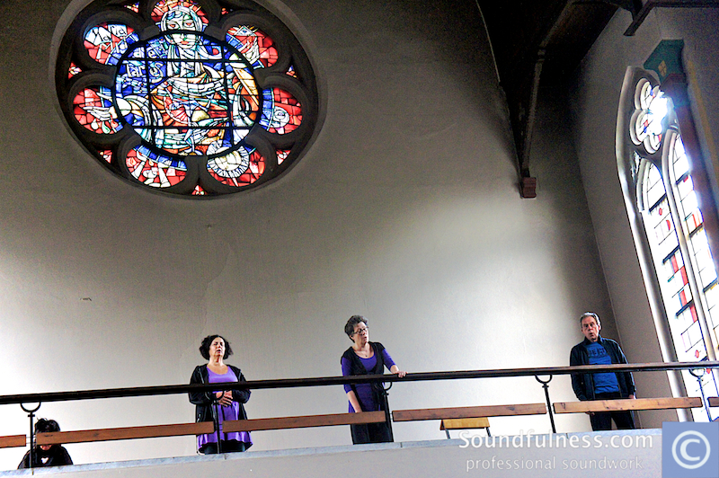 Three singers performing beside a stained-glass rose window in a church balcony