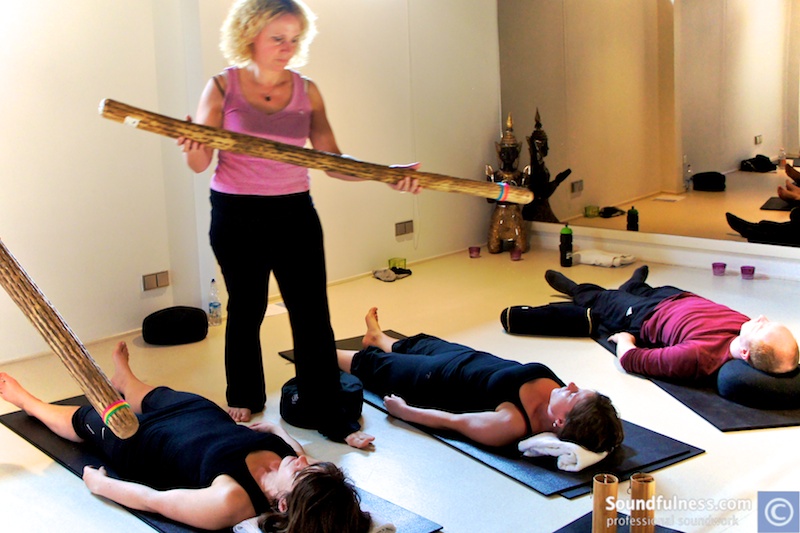 Practitioner passing a wooden rainstick over three clients lying on mats