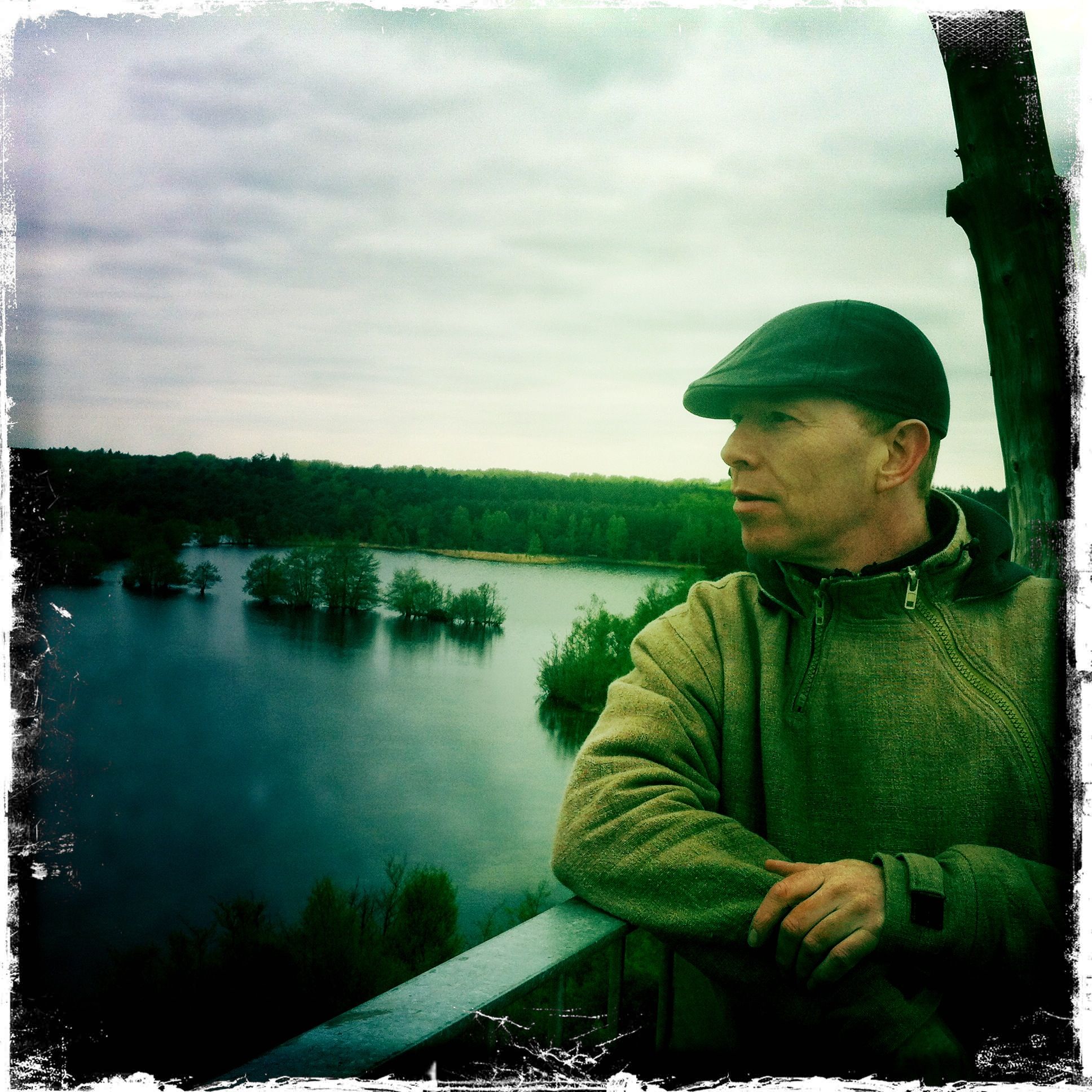 Coen Tuerlings in flat cap overlooking a still forest lake from a wooden tower