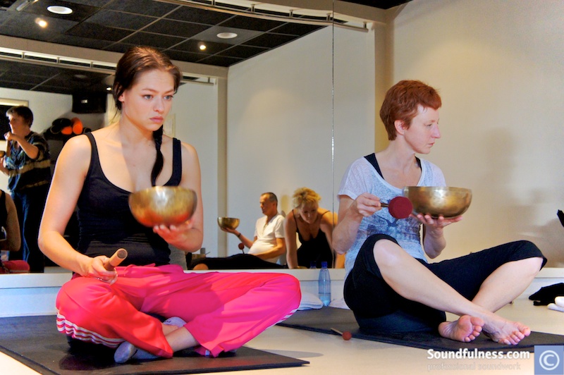 Two women seated on mats in a studio each holding a singing bowl and mallet