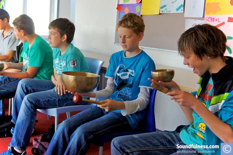 Row of boys in classroom seats each holding and playing their own singing bowl