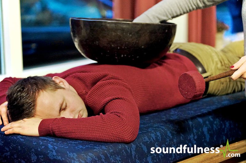 Client lying face down with a dark singing bowl resting on the lower back being struck with a mallet